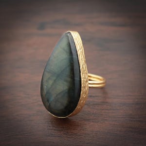 May include: A gold ring with a large, teardrop-shaped, dark green gemstone. The gemstone displays internal patterns. The ring's band and bezel have a textured, hammered appearance, creating a striking contrast in the design.