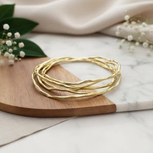 Puede incluir: Un conjunto de pulseras doradas con superficie texturizada. Las pulseras se exhiben sobre una superficie de madera y mármol, con flores blancas y hojas verdes en el fondo. Las pulseras son una joya sencilla y elegante.