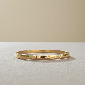 Puede incluir: Un brazalete dorado con una textura martillada. El brazalete es circular y parece estar hecho de metal. La superficie tiene un patrón texturizado e irregular. El fondo es de color neutro.