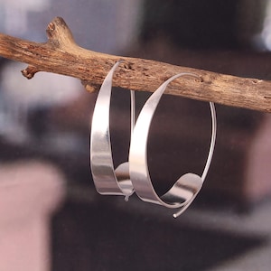 May include: A pair of silver hoop earrings with a unique, curved design. The earrings are suspended from a natural wooden branch, showcasing their modern, minimalist style. The polished metal reflects light, highlighting the smooth, flowing lines of the jewelry.