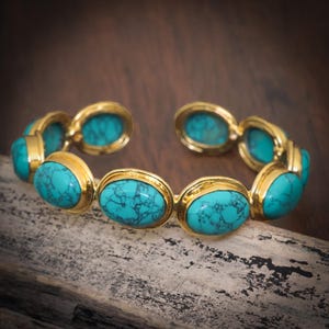 May include: Gold bracelet with oval turquoise stones. Each stone is set in a gold bezel. The bracelet is displayed on a wooden surface. The turquoise stones have a blue-green hue, and the gold setting adds a touch of elegance.