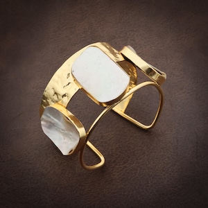 May include: A gold-toned cuff bracelet featuring a hammered metal finish and three irregular, smooth white shell inlays. The open-ended design consists of a wide band connected by thin, curved metal wires, creating a structured, layered appearance.