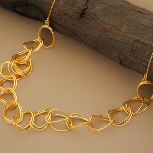 Puede incluir: Un collar dorado con una cadena de eslabones ovalados irregulares e interconectados. El collar se muestra sobre un fondo neutro con un elemento de madera marrón oscuro. La cadena es de metal brillante y reflectante.