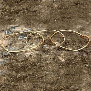 May include: Three delicate bangles in gold and silver tones rest on a textured surface. Two are gold, and one is silver. The bangles have a simple, minimalist design, perfect for everyday wear. The gold bangles have a hammered texture.