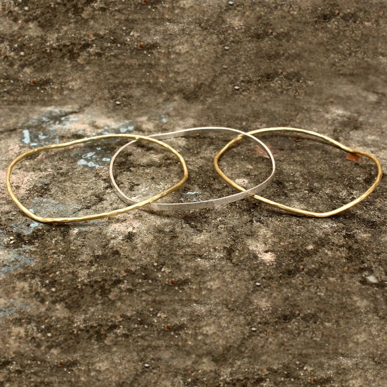 May include: Three delicate bangles in gold and silver tones rest on a textured surface. Two are gold, and one is silver. The bangles have a simple, minimalist design, perfect for everyday wear. The gold bangles have a hammered texture.