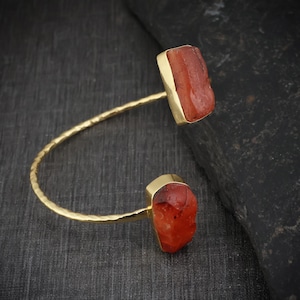 May include: A gold-toned open cuff bracelet featuring a textured, hammered metal band. The two ends of the bracelet are capped with raw, rectangular-shaped orange-red stones that have a translucent, crystalline appearance and a polished finish.