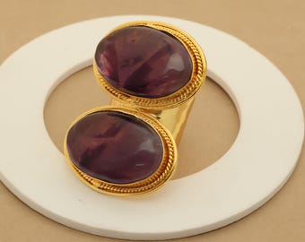 May include: A gold-toned ring featuring two oval, deep purple gemstones. The gemstones are set in a gold frame with a rope-like texture. The ring is displayed on a white, circular surface.