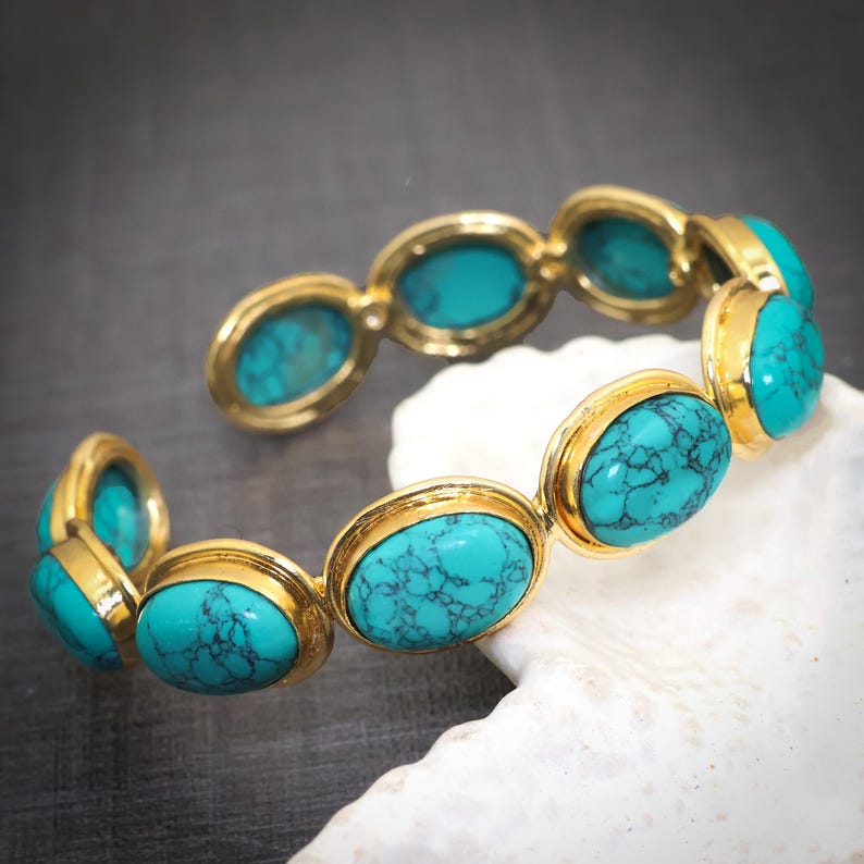 May include: Gold-coloured bracelet with oval turquoise stones. The hinged bracelet features turquoise stones set in gold-coloured bezels. The turquoise stones have a natural, veined appearance.