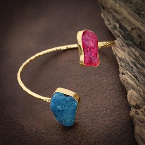 May include: A gold-toned open cuff bracelet featuring a hammered texture along the band. The bracelet is accented with two raw, irregular-shaped stones: one vibrant pink stone at one end and a deep blue stone at the other end of the opening.