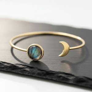 May include: A gold-colored bracelet featuring a round, iridescent stone and a crescent moon design. The stone displays shades of blue and green. The bracelet is set against a dark surface, highlighting its celestial theme.