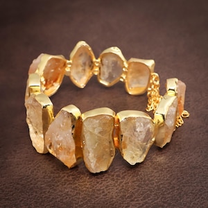 Könnte beinhalten: Ein Armband mit einer Reihe von rohen, durchscheinenden gelb-orangen Citrinsteinen, die jeweils in eine polierte goldfarbene Metallfassung eingefasst sind. Die Steine sind miteinander verbunden und das Stück hat eine feine goldfarbene Kette.