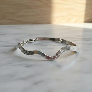 Könnte beinhalten: Ein silbernes Armband mit gehämmerten Texturen und einem wellenförmigen Design. Das Armband hat einen einfachen Verschluss und wird auf einer Marmoroberfläche präsentiert. Das Schmuckstück ist ein stilvolles Accessoire.