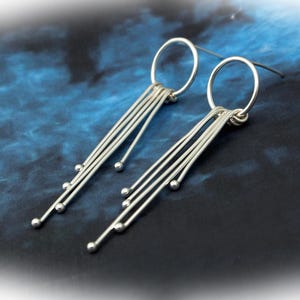 May include: A pair of silver dangle earrings. Each earring features a circular top connected to multiple straight, thin bars. Small silver spheres are attached to the ends of the bars, creating a linear, minimalist design. The earrings are displayed against a dark blue background.