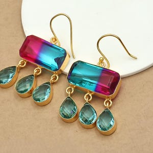 May include: Gold-coloured earrings with rectangular, colour-shifting stones in pink and blue. Each earring has three dangling, teal teardrop-shaped stones. The earrings are displayed against a neutral background.