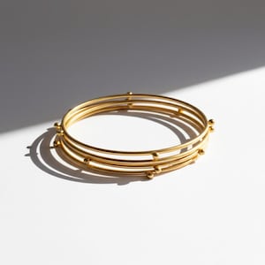 Könnte beinhalten: Ein Satz von vier goldfarbenen Armreifen. Jeder Armreif ist ein dünnes, glattes Band mit kleinen, gleichmäßig verteilten kugelförmigen Akzenten. Die Armreifen sind gestapelt und werfen einen Schatten auf eine weiße Oberfläche.