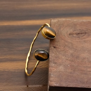 May include: A gold-toned cuff bracelet featuring two oval tiger's eye stones. The stones are set in gold bezels and the bracelet has a hammered texture. The bracelet is displayed on a wooden surface.