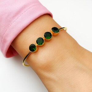 May include: A gold-toned bangle bracelet featuring four round, dark green gemstones. The bracelet is worn on a wrist, with a pink sleeve visible in the background. The gemstones are set in gold bezels.