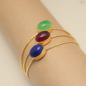 Puede incluir: Brazaletes dorados con tres piedras preciosas ovaladas en verde, rojo y azul. Las piedras están engastadas en biseles dorados y conectadas por tres bandas doradas texturizadas. Los brazaletes se llevan en una muñeca.