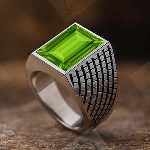 May include: A silver ring with a rectangular, bright green gemstone. The ring has a geometric pattern on the sides. The ring is set on a textured, brown surface.