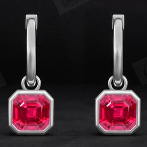 Puede incluir: Un par de pendientes de aro plateados con un colgante cuadrado de piedra preciosa roja brillante. Las piedras preciosas están engastadas en un bisel plateado, y los pendientes cuelgan de un aro sencillo. El fondo es oscuro.