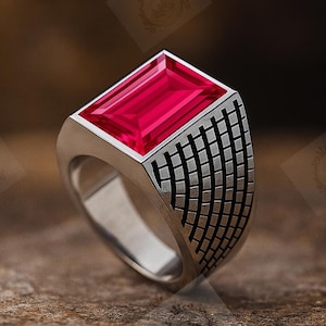 Könnte beinhalten: Ein silberner Ring mit einem rechteckigen, leuchtend roten Edelstein. Der Ring hat ein geometrisches Muster an den Seiten. Der Ring liegt auf einer strukturierten, braunen Oberfläche.