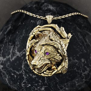 May include: A gold-coloured pendant necklace featuring a detailed wolf head with purple gemstone eyes. The wolf is set within an ornate frame, and the necklace is displayed on a dark, textured stone. The chain is also gold-coloured.