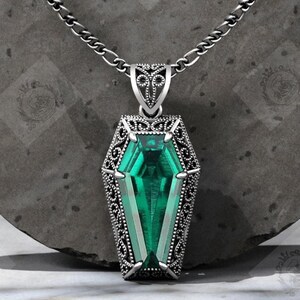 Puede incluir: Un collar con colgante plateado con una gema verde esmeralda en forma de ataúd. El colgante tiene intrincados detalles de filigrana y cuelga de una cadena. El diseño es gótico y elegante.