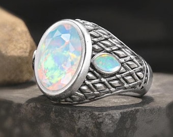 May include: A silver ring with an oval, iridescent opal gemstone as the centerpiece. The ring has a textured band with a smaller opal stone on the side. The opal displays a range of colors, including blue, green, and yellow.