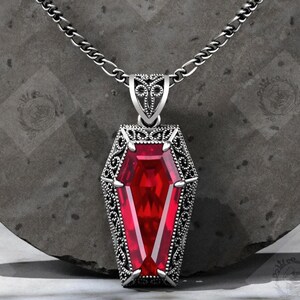 Op de afbeelding: Zilverkleurige hangerketting met een doodskistvormige hanger. De hanger heeft een grote, facetgeslepen rode edelsteen en gedetailleerde filigraan. De achtergrond is een donkergrijze, getextureerde ondergrond.