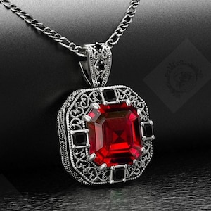 May include: A silver pendant necklace with a large, square, faceted red gemstone at its centre, framed by an ornate filigree design. The pendant is accented with black square stones and hangs from a silver chain. The background is black.