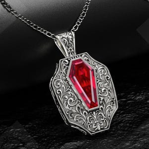 Könnte beinhalten: Silberfarbene Halskette mit Anhänger, der einen facettierten, roten Edelstein in Sargform aufweist. Der Anhänger ist in einem verzierten, gravierten Rahmen mit einer detaillierten Kette gefasst.