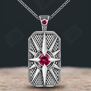 Könnte beinhalten: Silberfarbene Halskette mit Anhänger, rechteckigem Anhänger. Der Anhänger zeigt ein Kompassrosen-Design mit strukturiertem Hintergrund und einem zentralen roten Edelstein. Zusätzliche rote Edelsteine befinden sich oben. Die Kette ist silberfarben.
