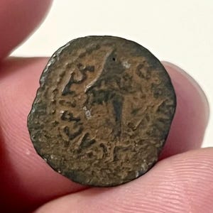 Può includere: Una moneta brunastra e usurata con testo visibile e un'immagine parziale. La superficie della moneta mostra segni di età e usura, con una forma leggermente irregolare. I dettagli suggeriscono che si tratta di un manufatto antico o storico.