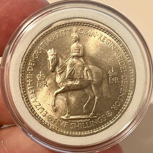 Op de afbeelding: Een zilverkleurige herdenkingsmunt in een doorzichtige plastic capsule. De voorzijde toont een figuur te paard, de tekst ELIZABETH II DEI GRATIA BRITT OMN REGINA FIDEI DEFENSOR en FIVE SHILLINGS, met twee kronen en de initialen ER.