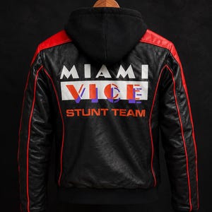 May include: A black faux leather jacket with a black hood, featuring red shoulder and sleeve accents. The back displays the white text MIAMI, the word VICE in orange and purple, and the orange text STUNT TEAM printed on a white rectangular panel.