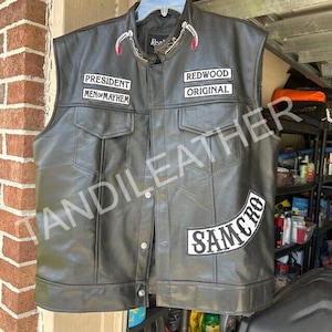 May include: A black leather vest featuring white embroidered patches. The chest patches read PRESIDENT, MEN OF MAYHEM, REDWOOD, and ORIGINAL. A large curved patch on the lower front reads SAMCRO. The vest has a snap-button front closure and two chest pockets.