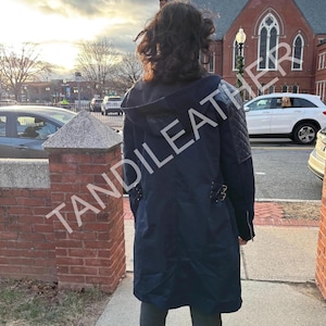 May include: A navy blue long coat with a hood and quilted shoulder detail. The coat has decorative buckles on the back and the text "TANDILEATHER" is visible on the back of the coat. The person is standing on a sidewalk.