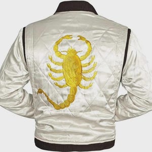 Puede incluir: Chaqueta de satén color crema con un diseño acolchado. La espalda presenta un gran escorpión bordado en oro. La chaqueta tiene cuello, ribetes y puños negros. Una prenda de abrigo elegante.