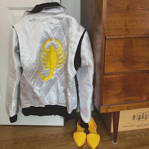 Könnte beinhalten: Eine gesteppte silberne Satin-Bomberjacke mit einem großen, gestickten gelben Skorpion auf der Rückseite. Die Jacke hat einen schwarzen Kragen, schwarze Ärmelbündchen und einen schwarzen Bund. Ein Paar gelbe spitze Ballerinas steht unter der Jacke.
