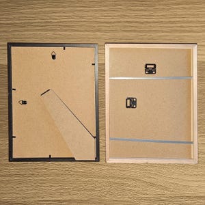 Puede incluir: Se muestran dos marcos de fotos rectangulares, uno con borde negro y otro con borde de madera clara. Se ven los respaldos de los marcos, mostrando respaldo de cart&oacute;n, herrajes para colgar y un soporte tipo caballete en el marco negro.