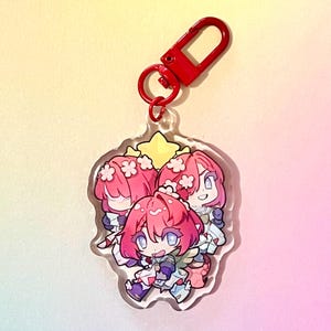 Puede incluir: Un llavero de acr&iacute;lico transparente con una ilustraci&oacute;n de dibujos animados de tres personajes con cabello rosado, ropa blanca y detalles de flores. El llavero tiene un cierre de metal rojo.