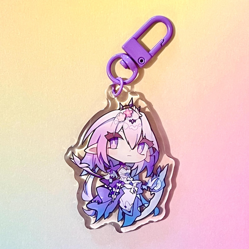 Puede incluir: Un llavero de un personaje de anime morado y azul con un clip morado. El personaje tiene el pelo largo rosa, un vestido blanco y una cola de drag&oacute;n azul y blanca.