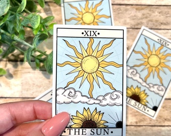 Puede incluir: La carta del tarot El Sol presenta un sol amarillo con rayos, un girasol y nubes sobre un fondo azul. El número de la carta XIX está impreso en negro en la parte superior y "EL SOL" está impreso en negro en la parte inferior.