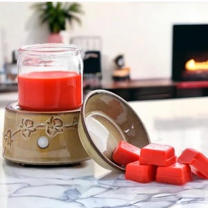 May include: A ceramic wax warmer with a red candle in a glass jar, and several red wax cubes. The warmer is tan with a floral design. The items are on a white marble surface.