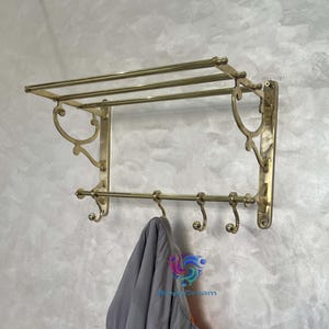 Puede incluir: Un perchero de pared de latón con una balda superior y varios ganchos. Una chaqueta gris y naranja cuelga de los ganchos. El perchero está montado en una pared gris texturizada.