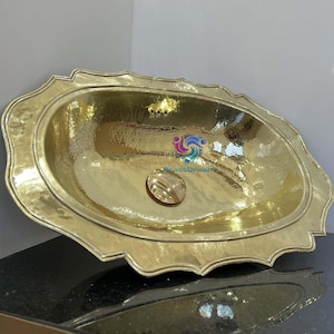 Puede incluir: Un lavabo ovalado de latón martillado con un borde festoneado decorativo. El lavabo tiene un acabado pulido y un desagüe central. El lavabo se exhibe sobre una encimera negra.