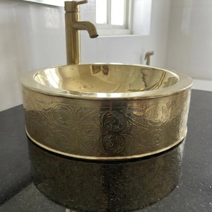Peut inclure: Un lavabo de salle de bain rond de couleur or avec un motif floral orné gravé sur l'extérieur. Un robinet doré assorti est monté au-dessus de l'évier. L'évier est posé sur un comptoir sombre.