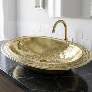 Pode incluir: Uma pia de banheiro oval dourada martelada com uma borda floral ornamentada. A pia está sobre uma bancada de mármore escuro, com uma torneira dourada. É um acessório de banheiro de luxo.