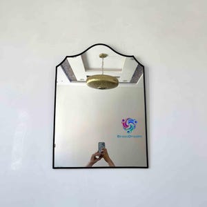 May include: A rectangular mirror with a black frame and a unique, scalloped top edge. The mirror reflects a room with a ceiling light fixture and the text "BrassDream". The mirror is mounted on a white wall.