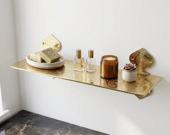 May include: A gold-colored metal shelf with a decorative spade design, holding a small white dish with green soap, two perfume bottles, a candle in a brown jar, and a small potted succulent. The shelf is mounted on a white wall.